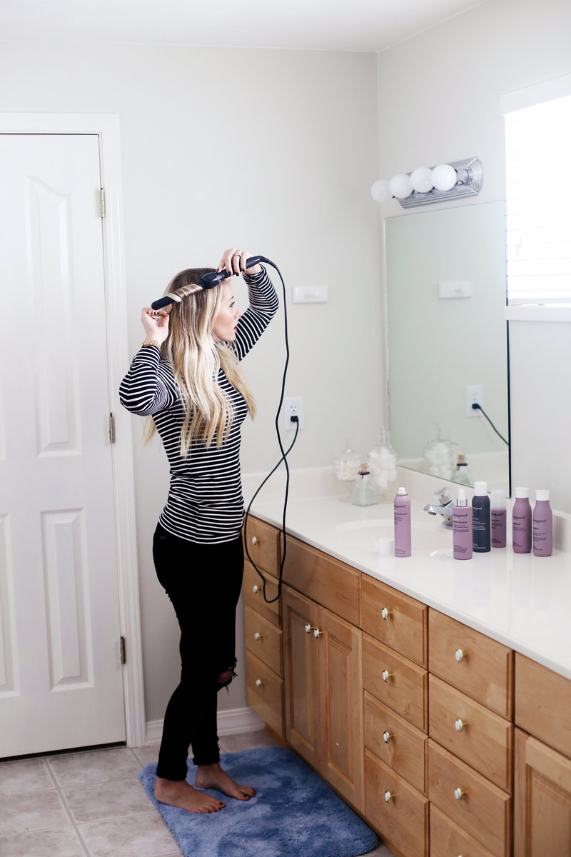 Abby stands in a white bathroom, using a curling iron on her hair.
